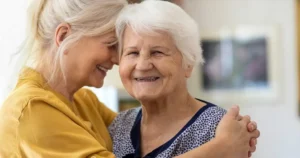 Eine lächelnde ältere Frau mit weißem Haar wird von einer jüngeren Frau mit blondem Haar umarmt. Beide wirken in einem warm beleuchteten Innenraum glücklich und verbunden und spiegeln die mitfühlende Unterstützung von 24 Stunden Pflege durch das Deutsche Pflegehilfswerk wider.