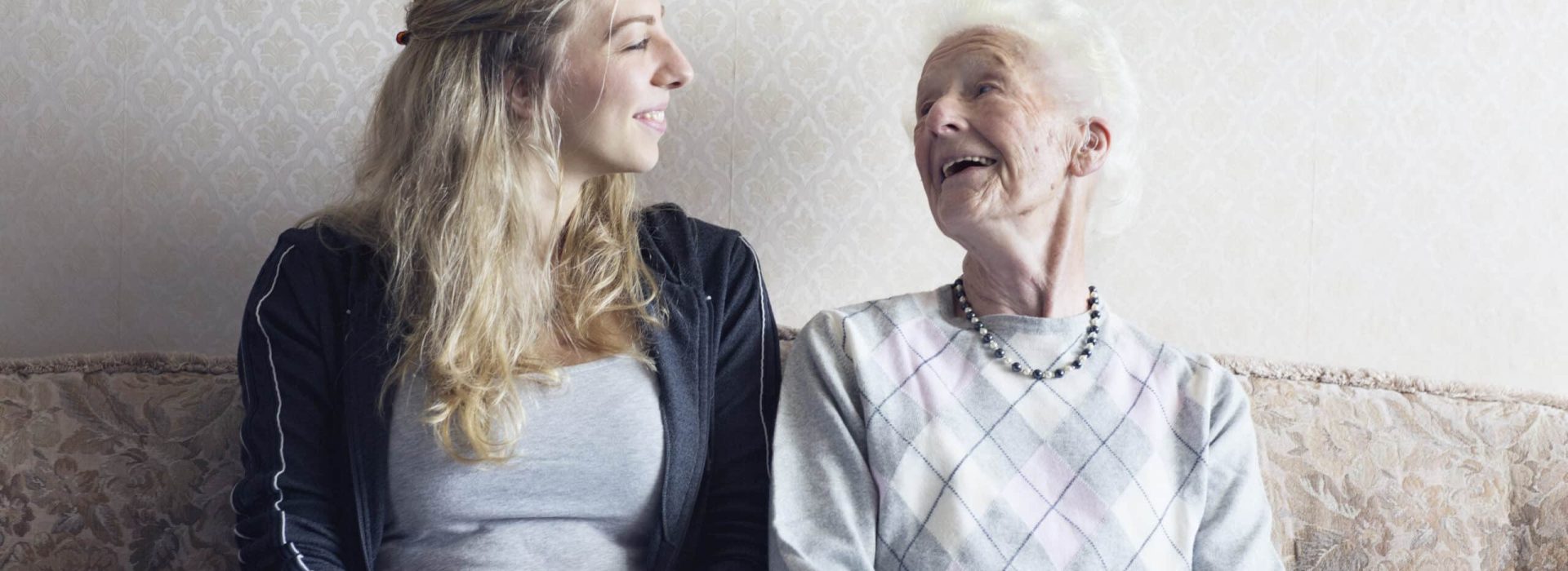 Eine Betreuerin sitzt mit einer 90-jährigen Frau auf einem Sofa, ist in ein Gespräch vertieft und teilt eine herzliche Bindung.